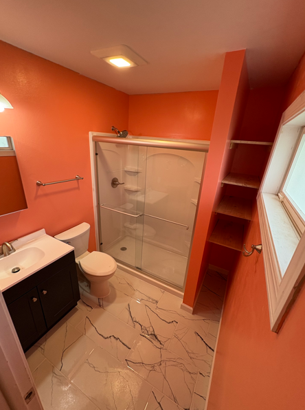 Small orange bathroom with glass shower, toilet, sink vanity, and a window.