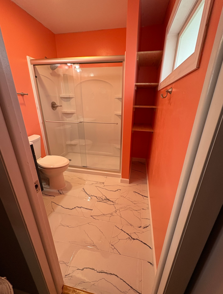 Orange bathroom with a tiled shower, toilet, marble floor, and window shelves