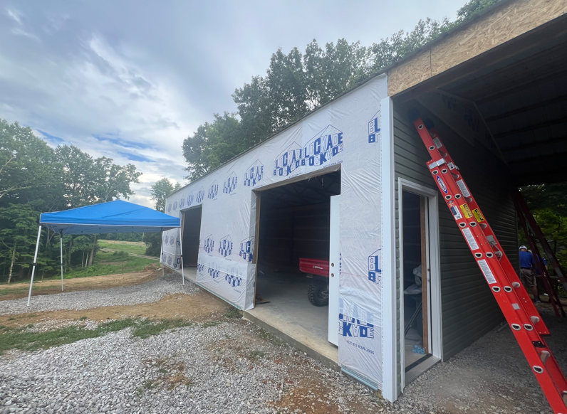 Partially built white garage with open bays, red ladder, and blue canopy on gravel driveway