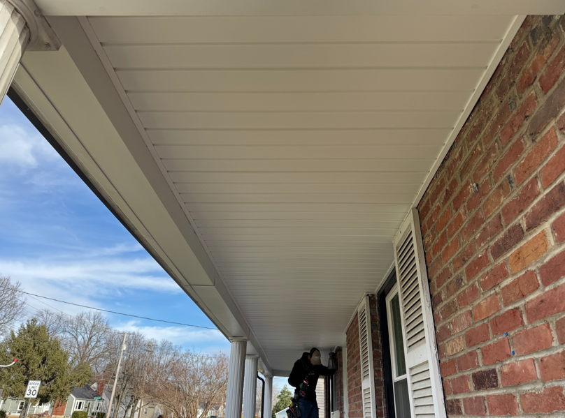 Person on a brick house porch under a white soffit, reaching toward a window near a white shutter.