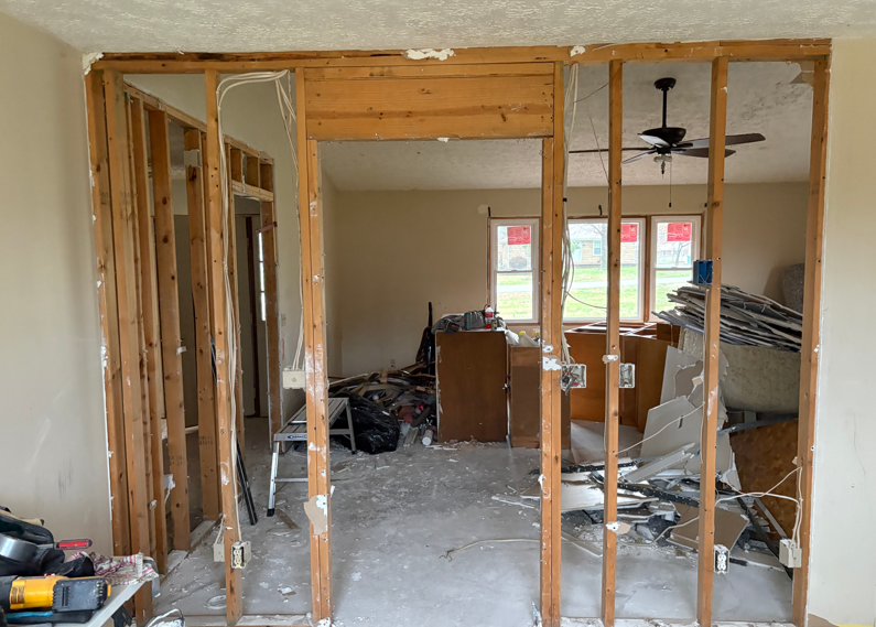 Partially demolished room with exposed studs, debris, and a ceiling fan visible through the opening