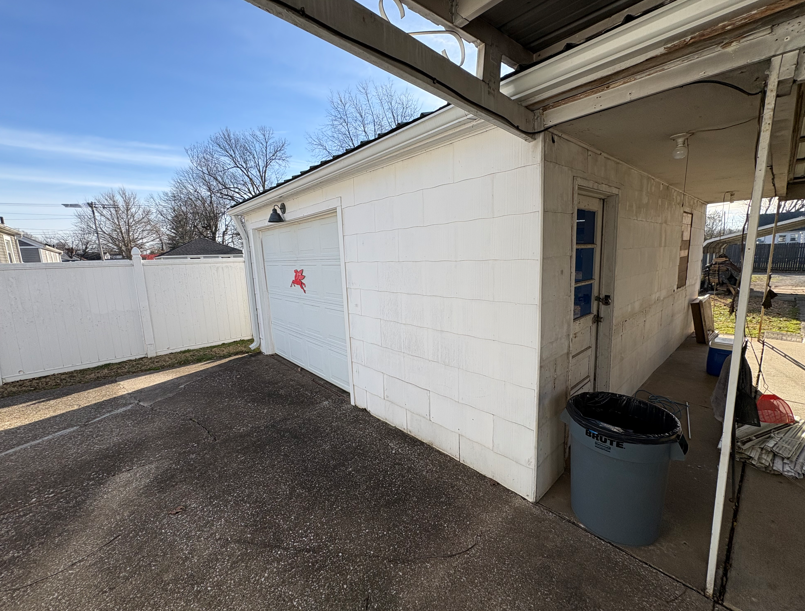Side yard beside a white building with a garage door, trash can, and covered patio.