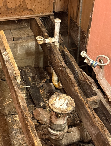 Rotted floor framing around exposed plumbing pipes and a drain opening in a damaged bathroom area