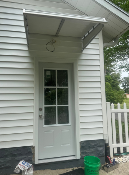 Back door entrance with white siding, awning, and a green bucket beside the doorway