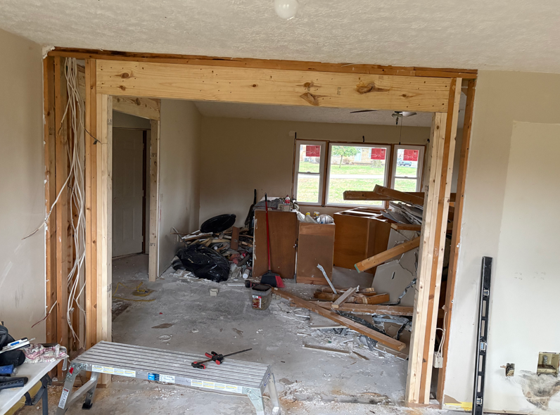 Interior renovation with exposed framing, debris on floor, and partially removed wall opening into another room