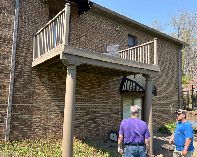 Two people standing beside a raised balcony and support columns on a brick building.