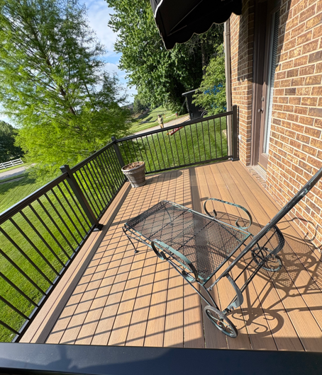Brick patio balcony with black railings, glass table, chairs, and a potted plant overlooking a green yard