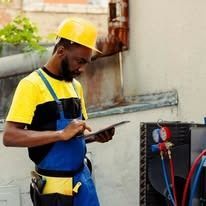 Black man in hardhat and work clothes using a tablet outdoors, near equipment.