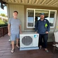 Two men pose with an air conditioning unit outdoors. One man in blue shirt, another in a polo.