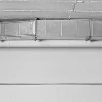 Metal HVAC ductwork installed on a white wall, below a cracked concrete ceiling.