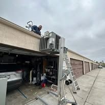 Man on roof, lifting HVAC unit. Ladder, garage, and storage units. Cloudy day.