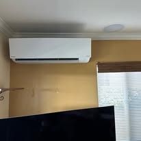 White ductless air conditioner on a gold wall, with a television screen and window visible.
