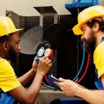 Two men in yellow shirts and hard hats working on an air conditioning unit.