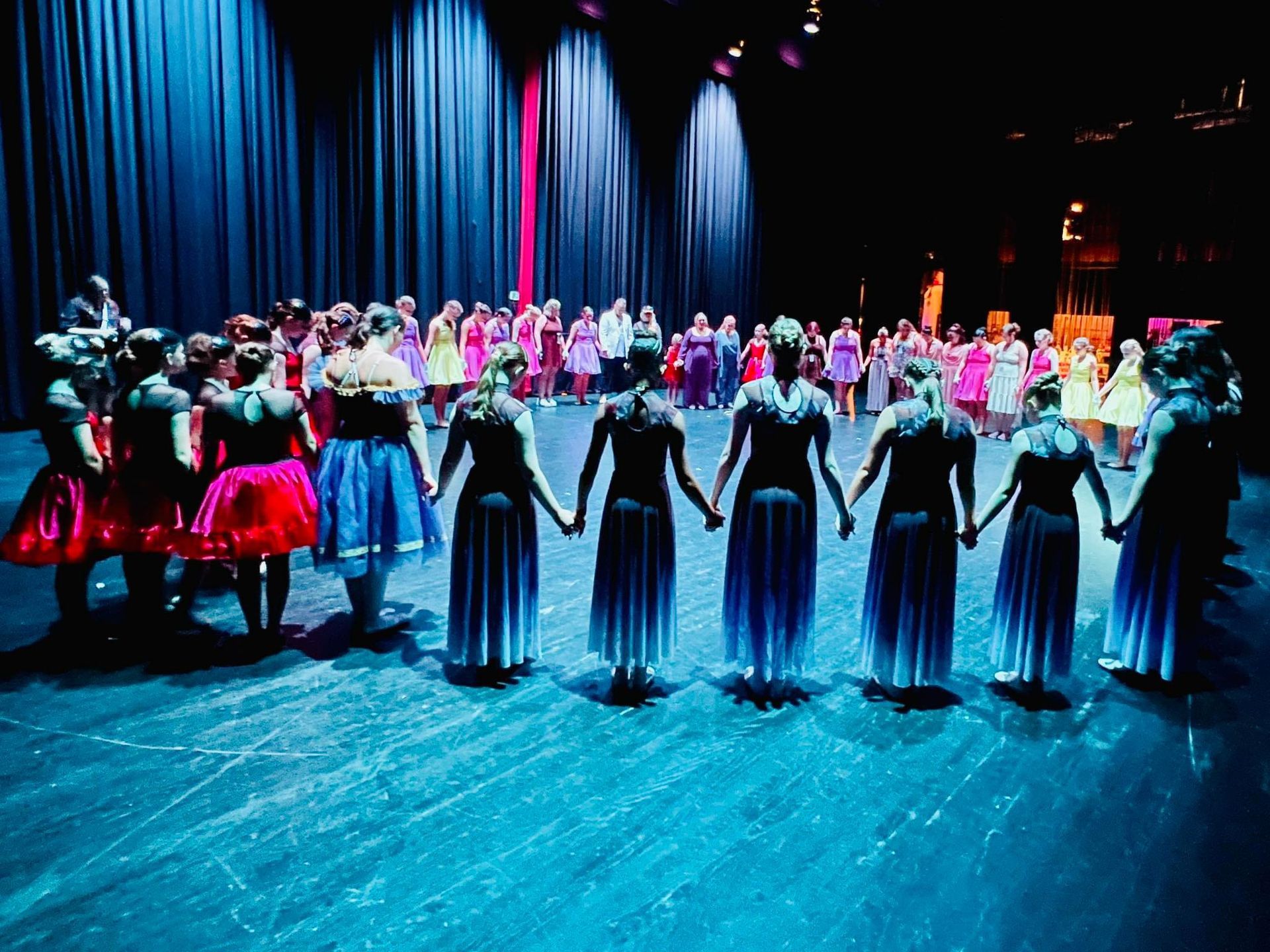 Dancers in colorful costumes stand in a circle on a stage, holding hands, with a dark curtain backdrop.