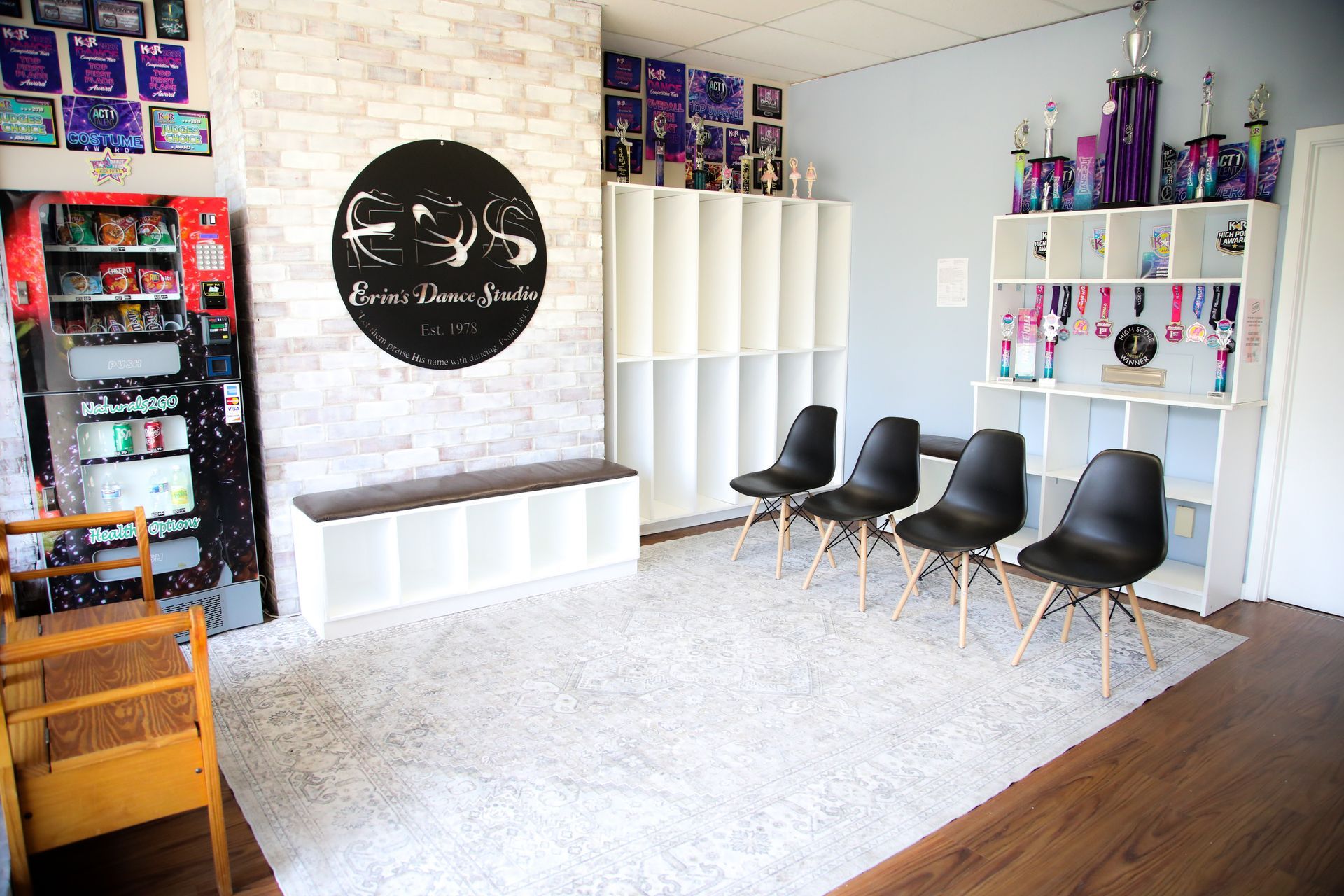 Dance studio waiting area with black chairs, white shelves with trophies, and a snack vending machine.