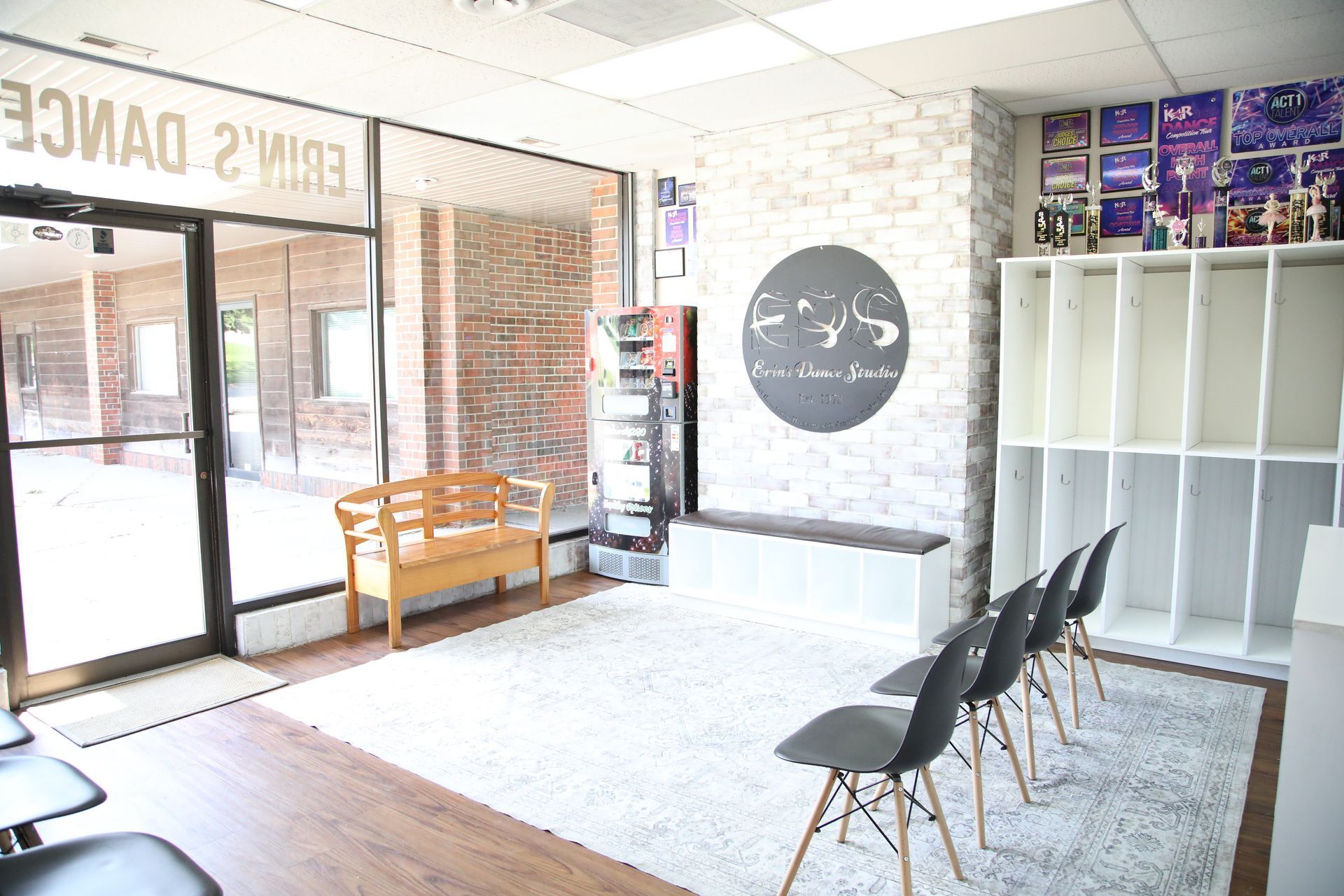 Dance studio lobby with bench, chairs, a white brick wall, and a large window.