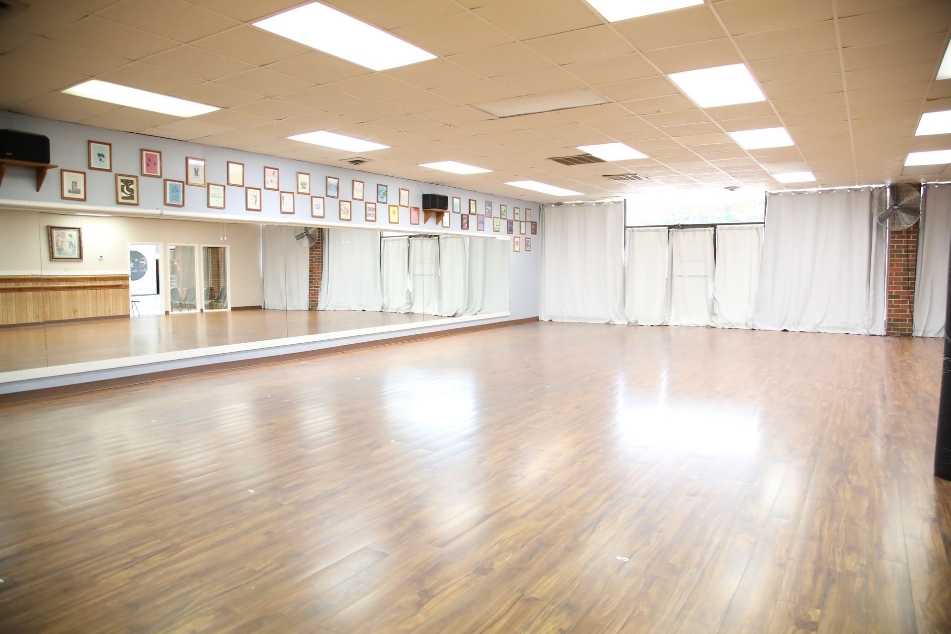 Dance studio with wood floor, mirrored wall, framed pictures, and white curtains.