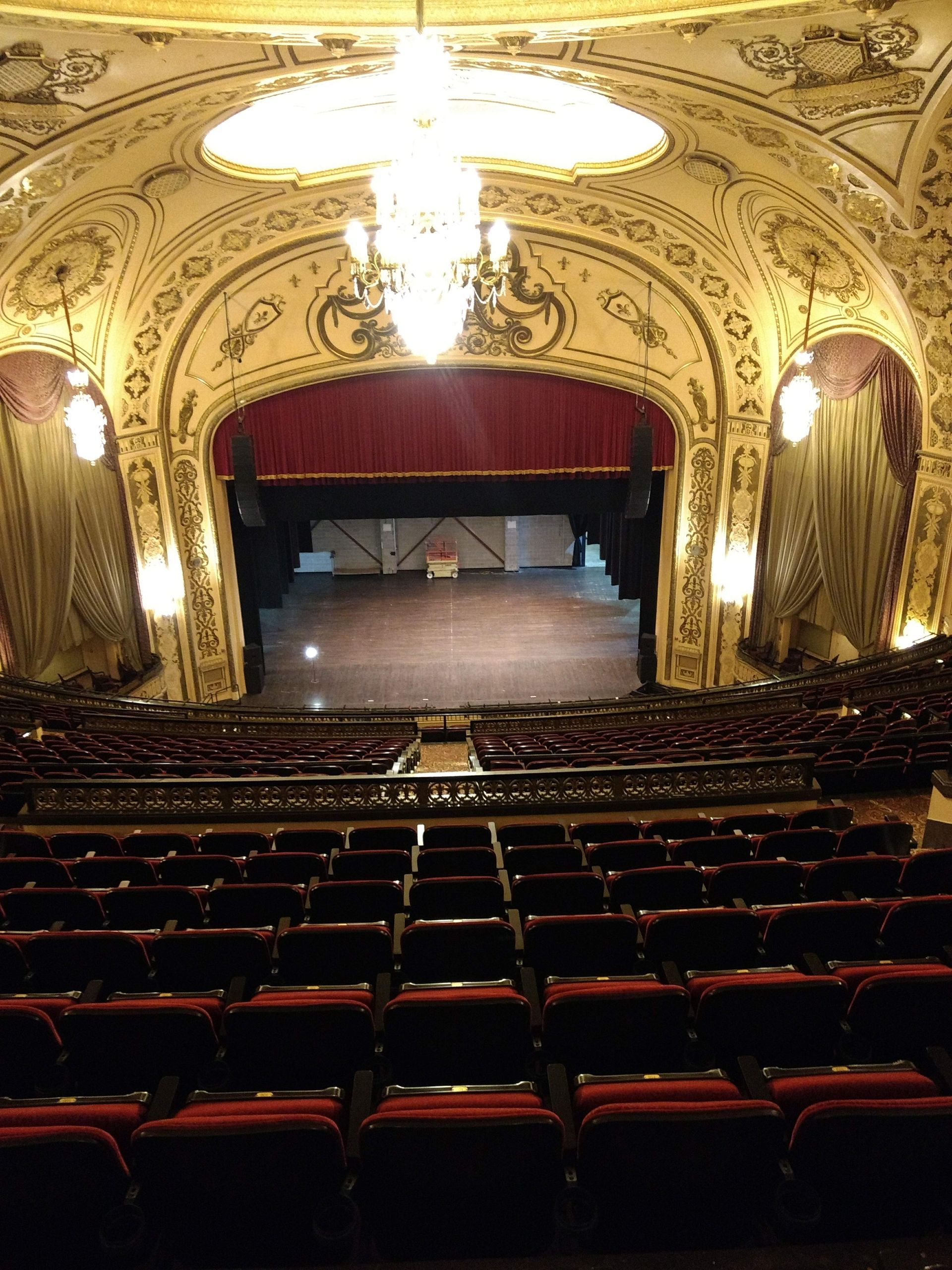 A grand theater interior. Red velvet seats face a stage with a closed red curtain. Ornate gold trim adorns the walls and ceiling.