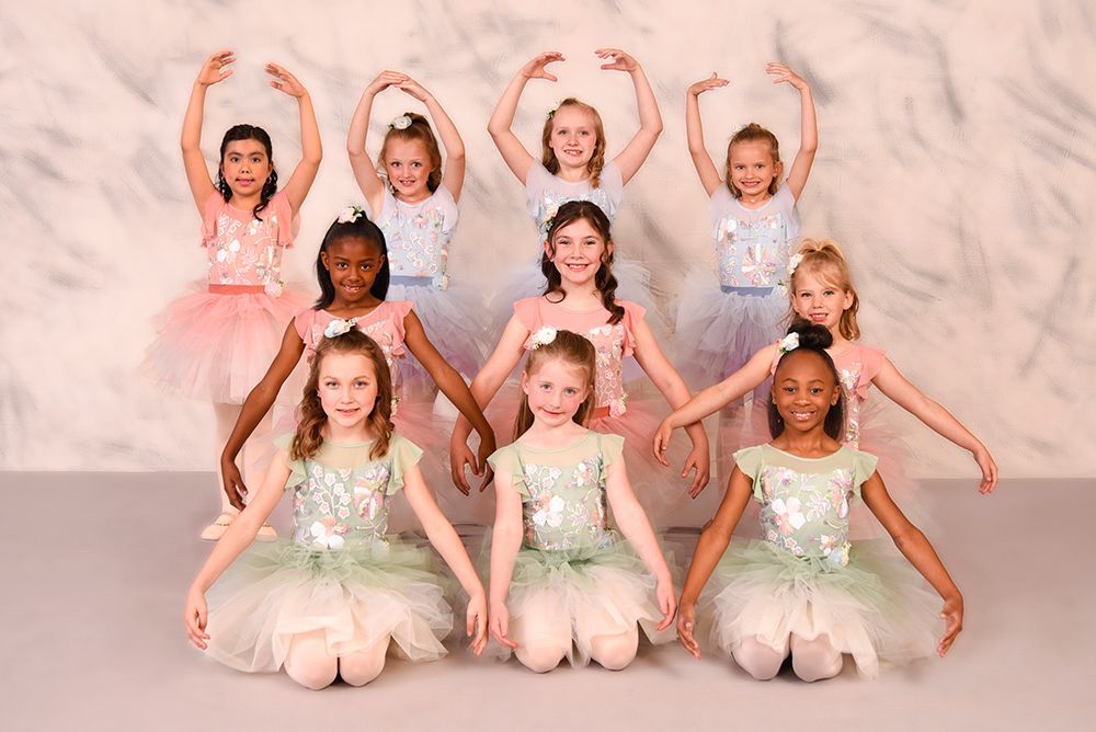 A group of young girls in ballet tutus poses. They are in a studio with arms gracefully overhead and out to the sides.