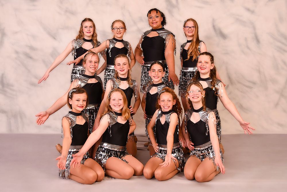 A group of young dancers in matching silver and black costumes strike poses in front of a cloudy backdrop.