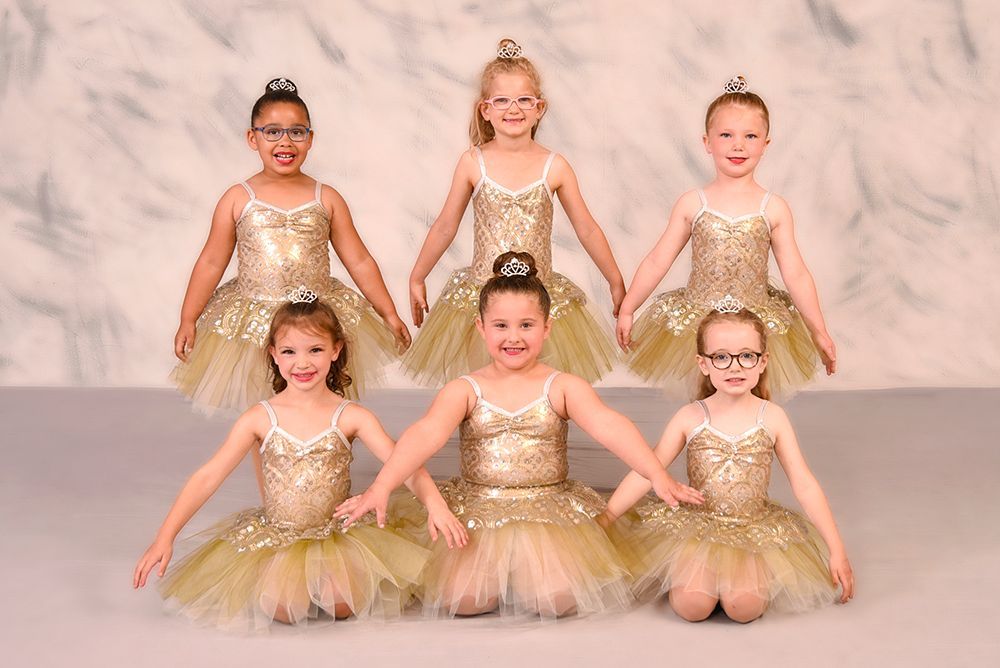 Six young ballerinas in gold tutus and tiaras, posing and smiling in a studio setting.