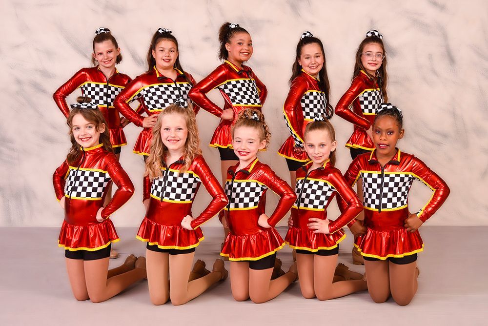A group of young girls in red, black, and yellow dance costumes pose for a photo against a neutral backdrop. They are smiling and have their hands on their hips.