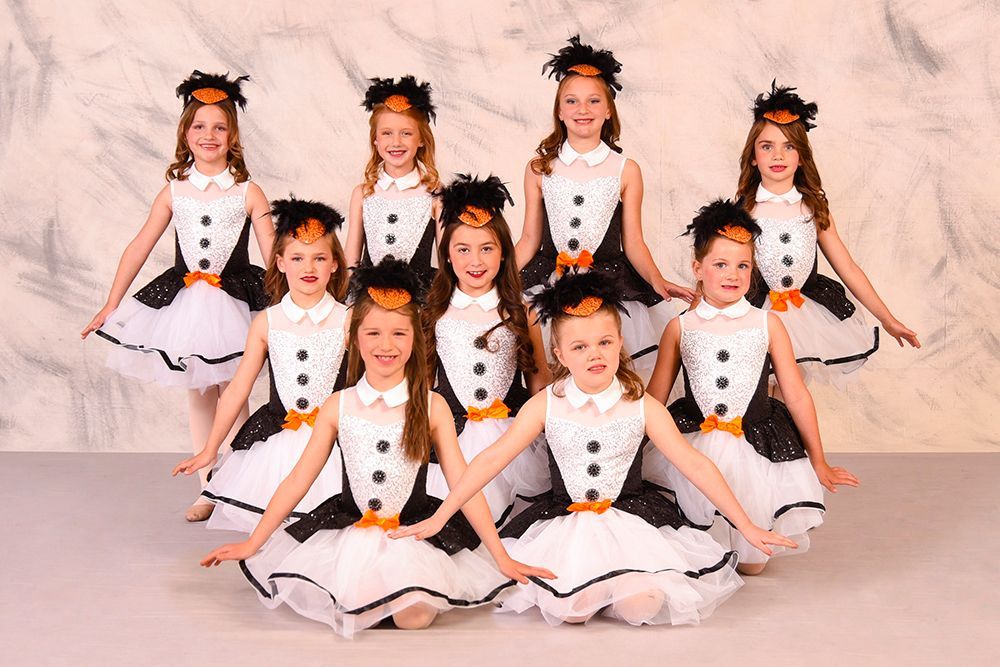 A group of young girls in penguin costumes with black and white outfits and feather headbands, posing in a studio.