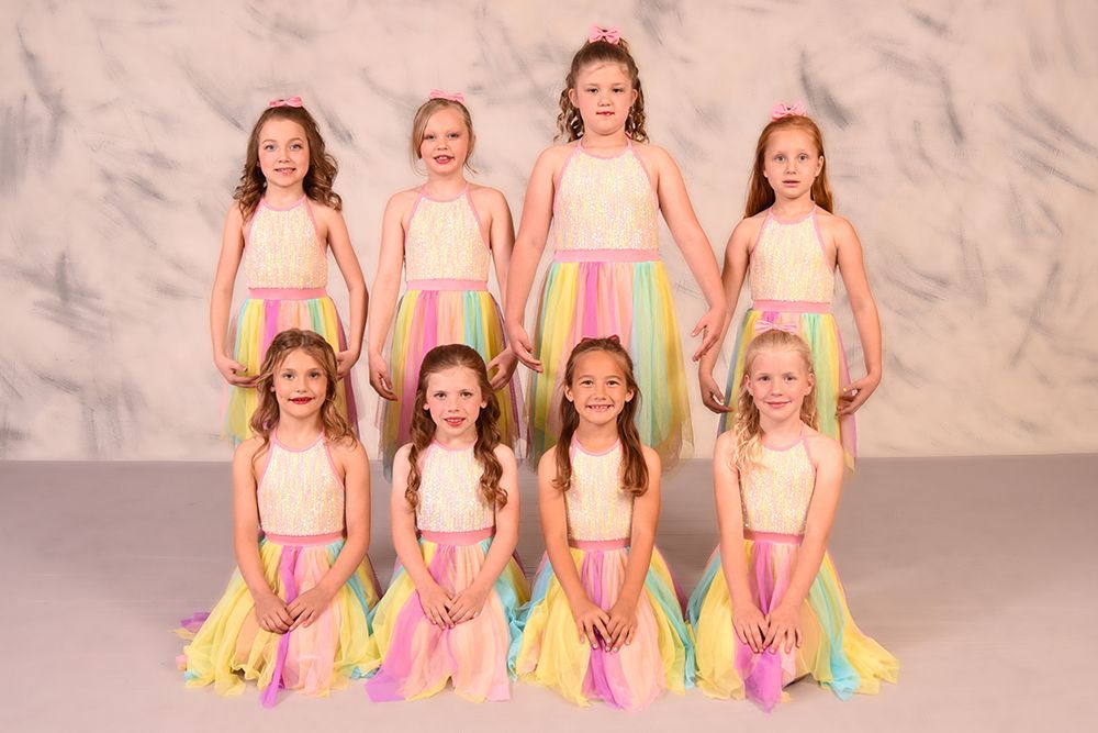 Group of young girls in colorful dresses posing for a photo in front of a neutral background. They're all smiling.