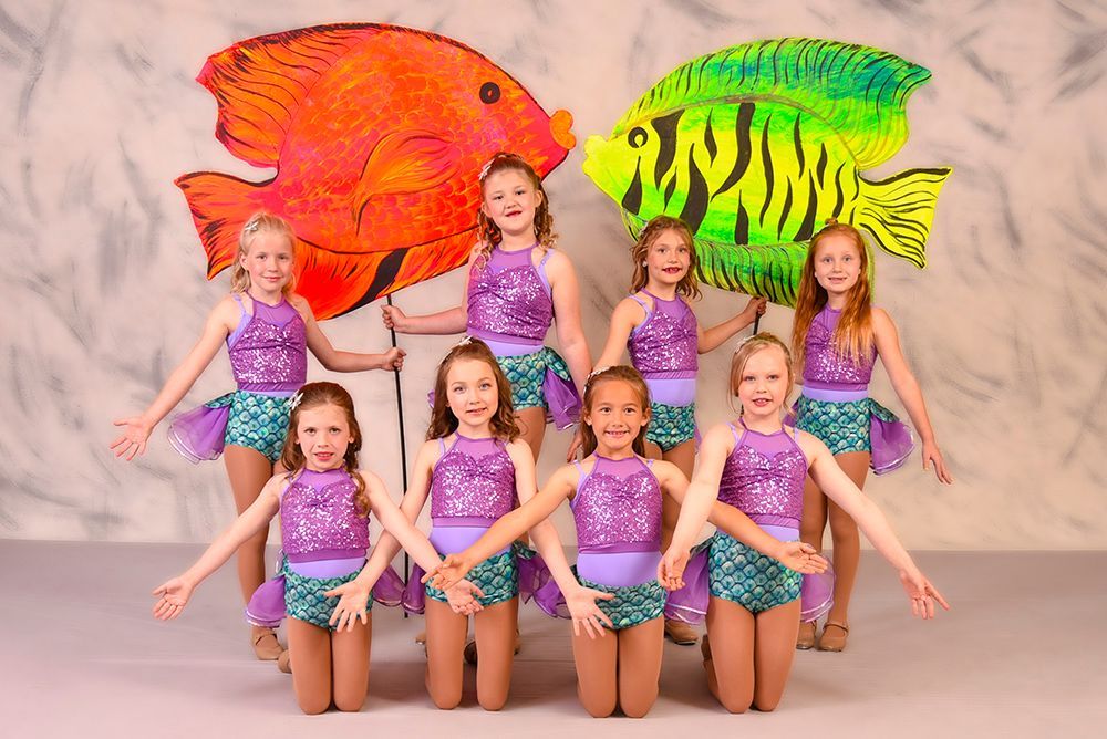 A group of young girls in mermaid costumes pose with large fish cutouts in front of a backdrop.