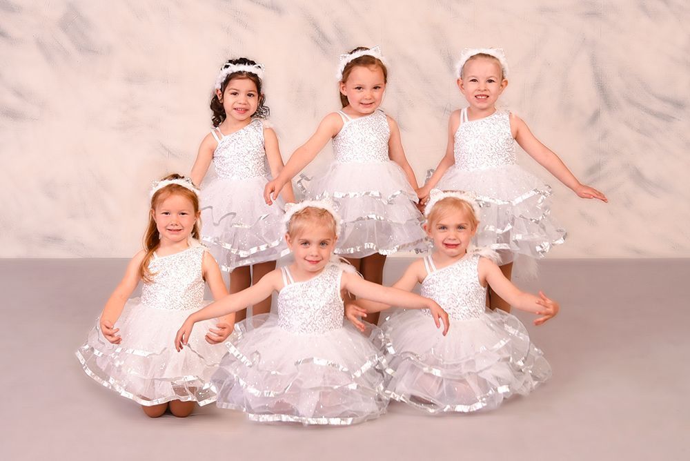 Six young girls in white dresses and headbands holding hands and smiling against a marbled backdrop.