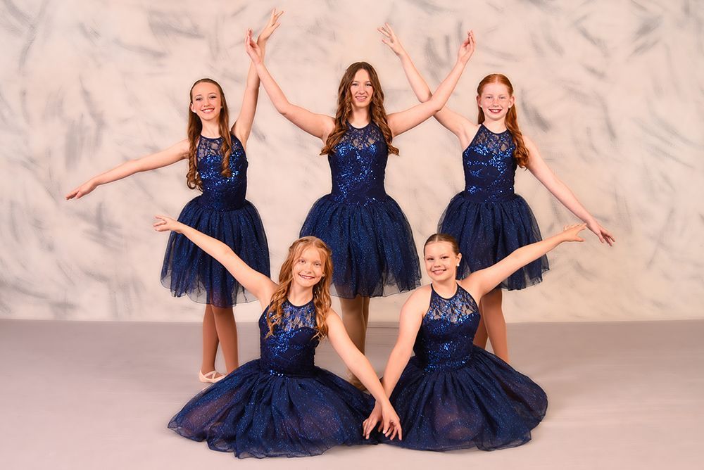 Five young dancers in navy blue dresses pose with arms outstretched against a mottled gray background.