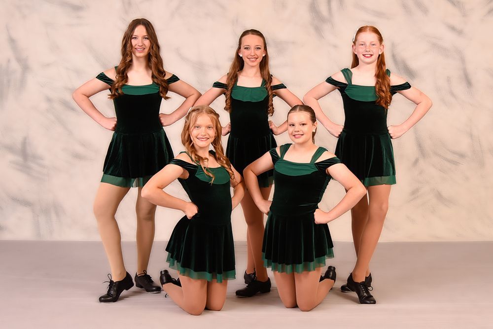 Five young female dancers in dark green dresses pose with hands on hips in front of a mottled gray background.