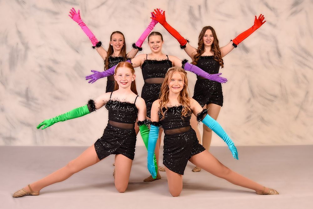 A dance team poses in a studio, wearing black sequined dresses and colorful gloves with their arms raised.