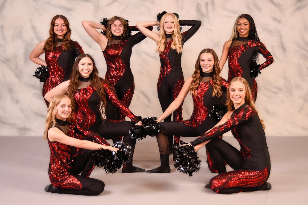 A cheerleading squad in black and red sequined uniforms poses in front of a swirl background, holding pom-poms.