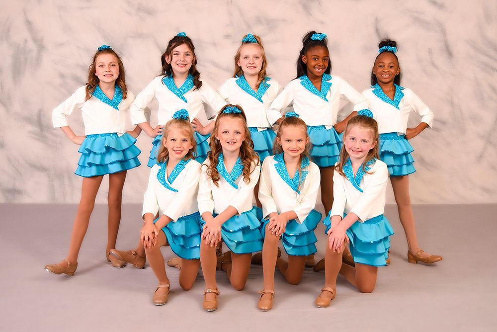 A group of young dancers in white and teal outfits pose on a studio backdrop.