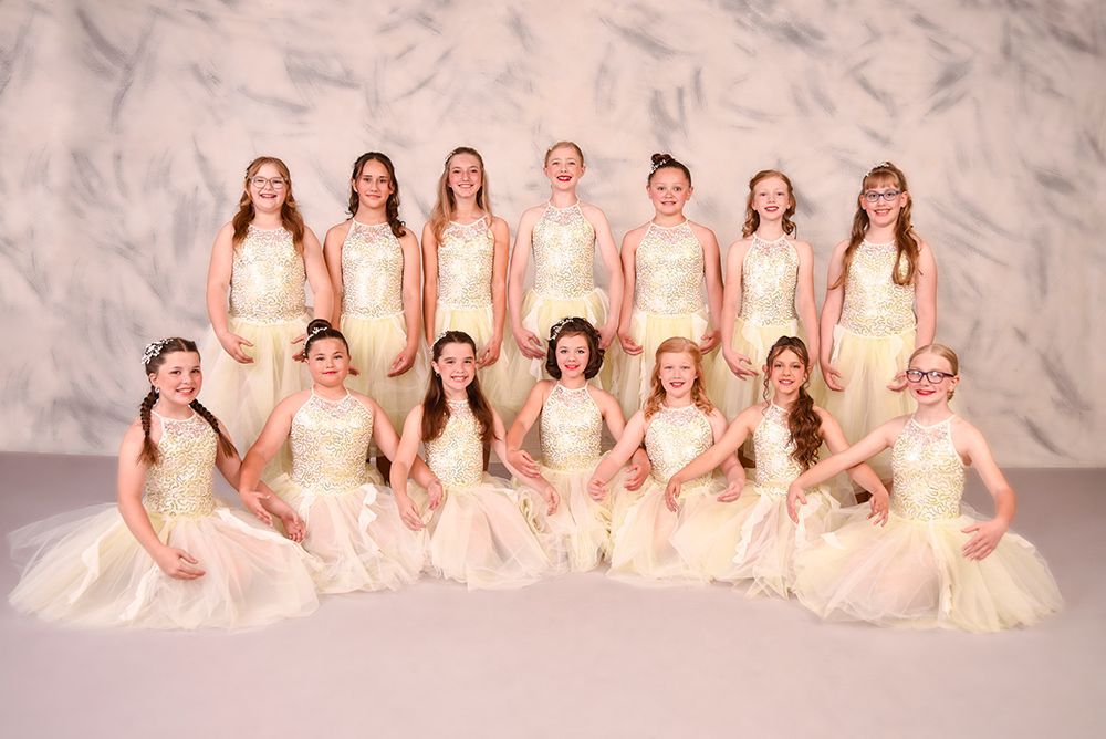 A group of young girls in cream-colored dresses pose for a studio photo. They are smiling, some kneeling and some standing.