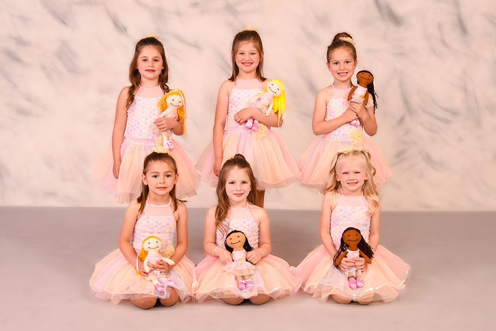 Six young girls in pink tutus pose with dolls against a painted background. They have smiling faces.