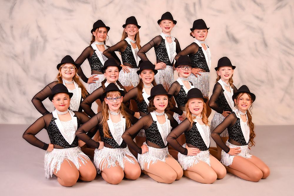 A group of young dancers in black and white costumes and hats, posing in a studio.