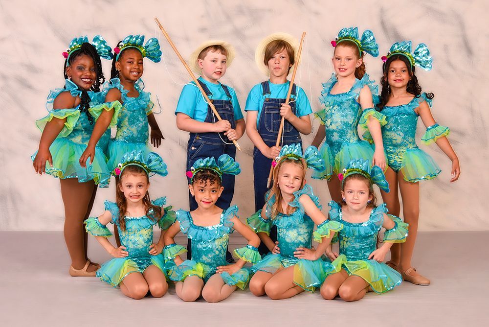 A group of children in turquoise costumes and two boys in overalls pose for a photo. The girls wear elaborate headpieces and dance attire.