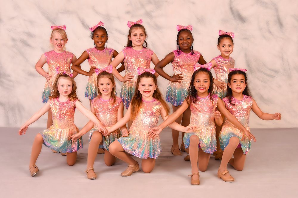 Ten young dancers in matching pink and rainbow sequined costumes pose in front of a blurred, light-colored backdrop.