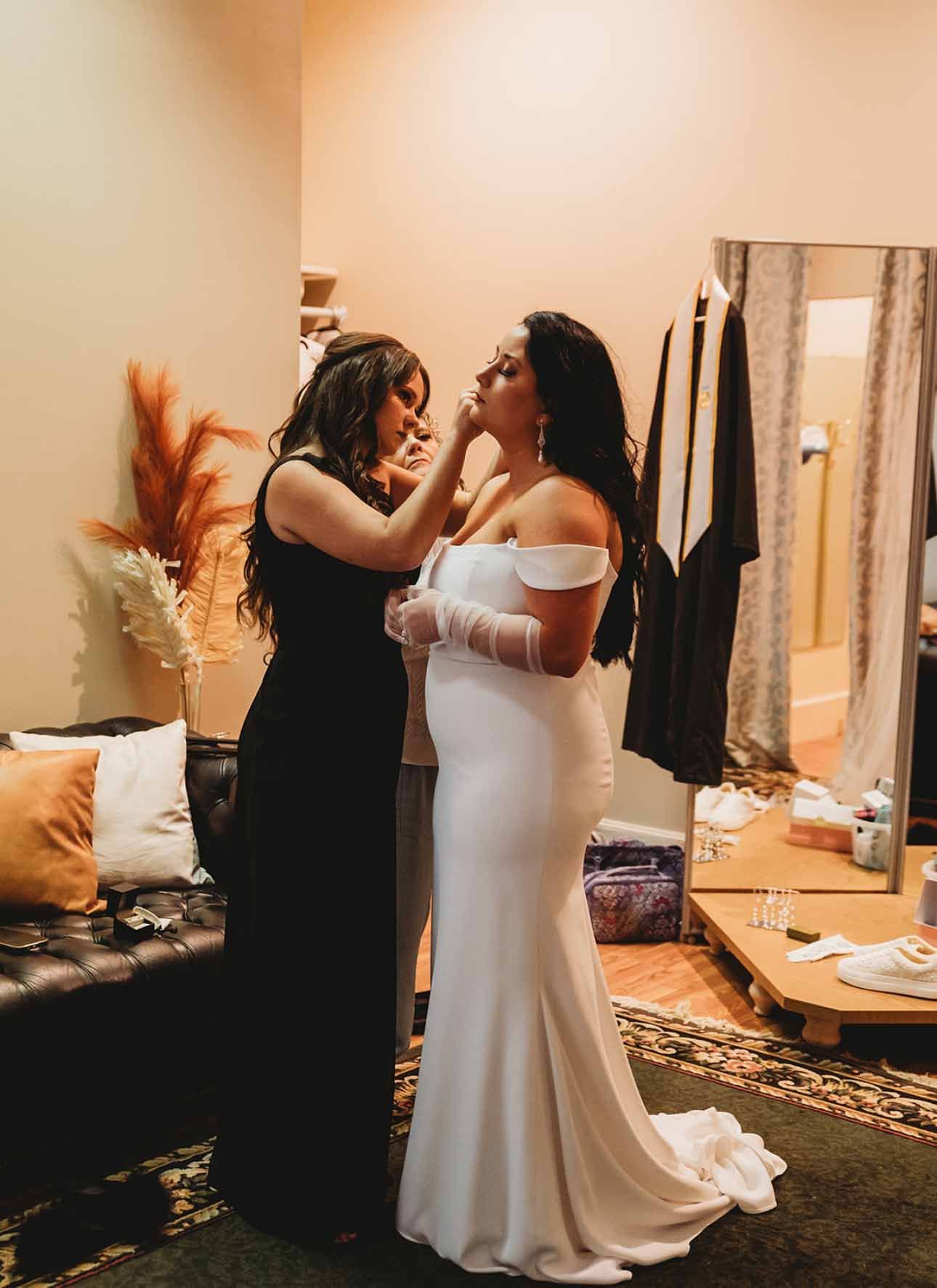 A woman in a white dress is getting her makeup done by a woman in a black dress.