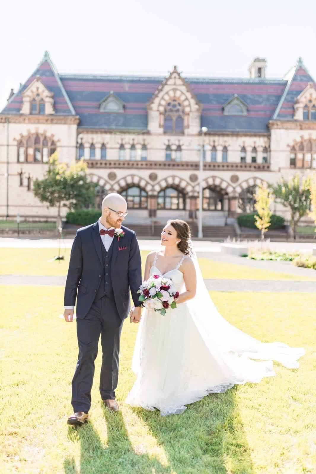 A bride and groom are walking in front of a large building holding hands.