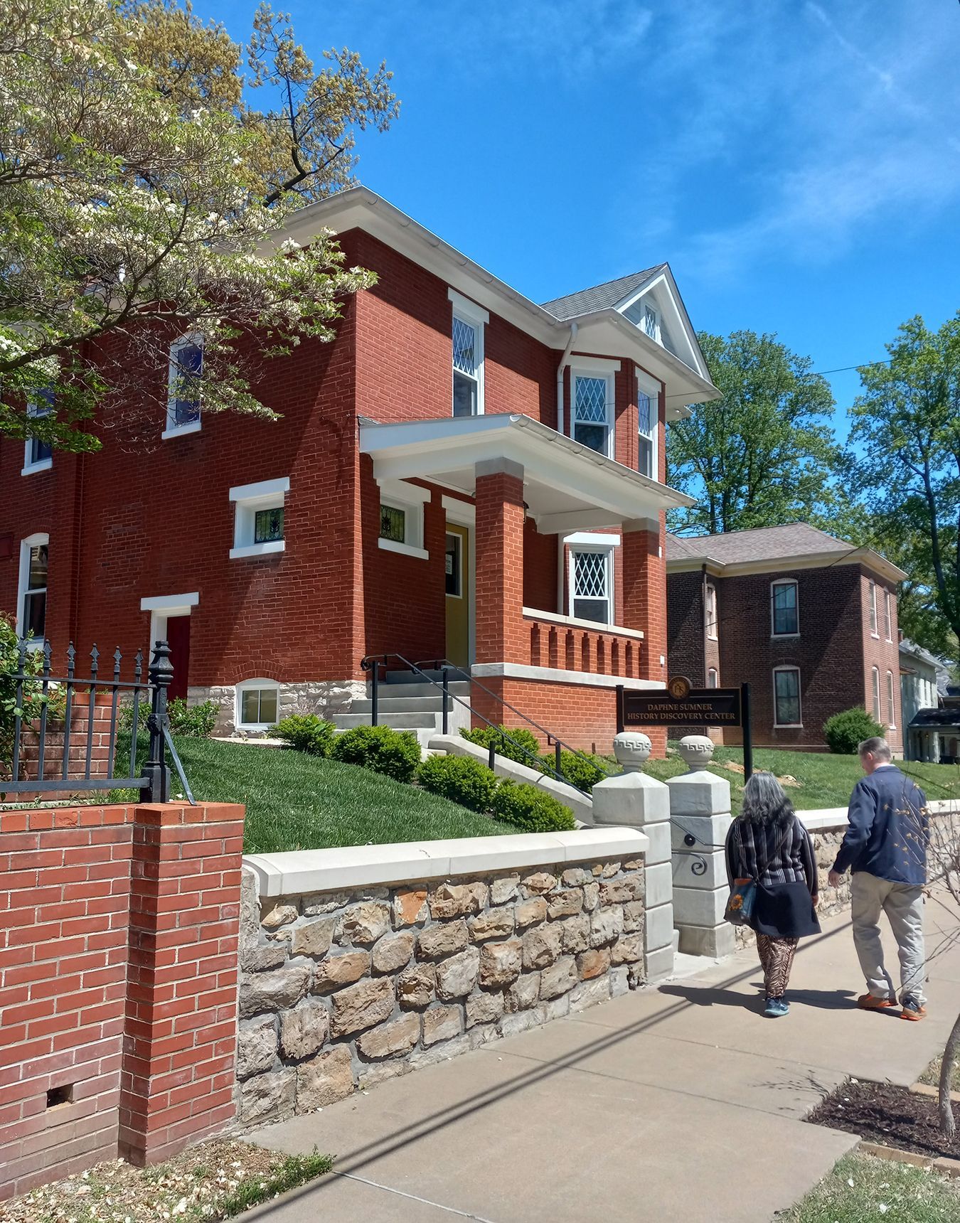 Two people are walking down a sidewalk in front of a large red brick house.