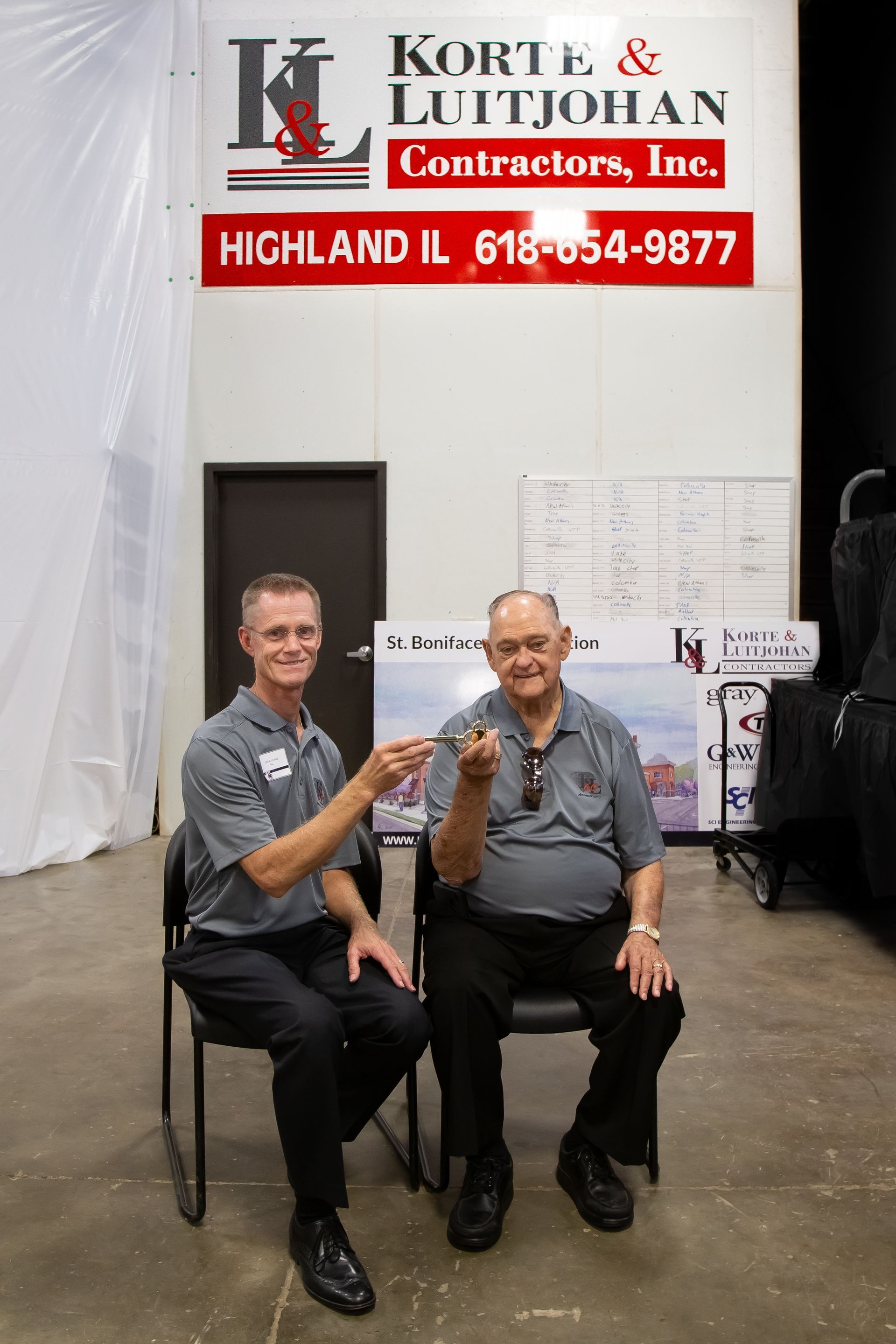Two men are sitting in chairs in front of a sign for korte & littjohan contractors inc.