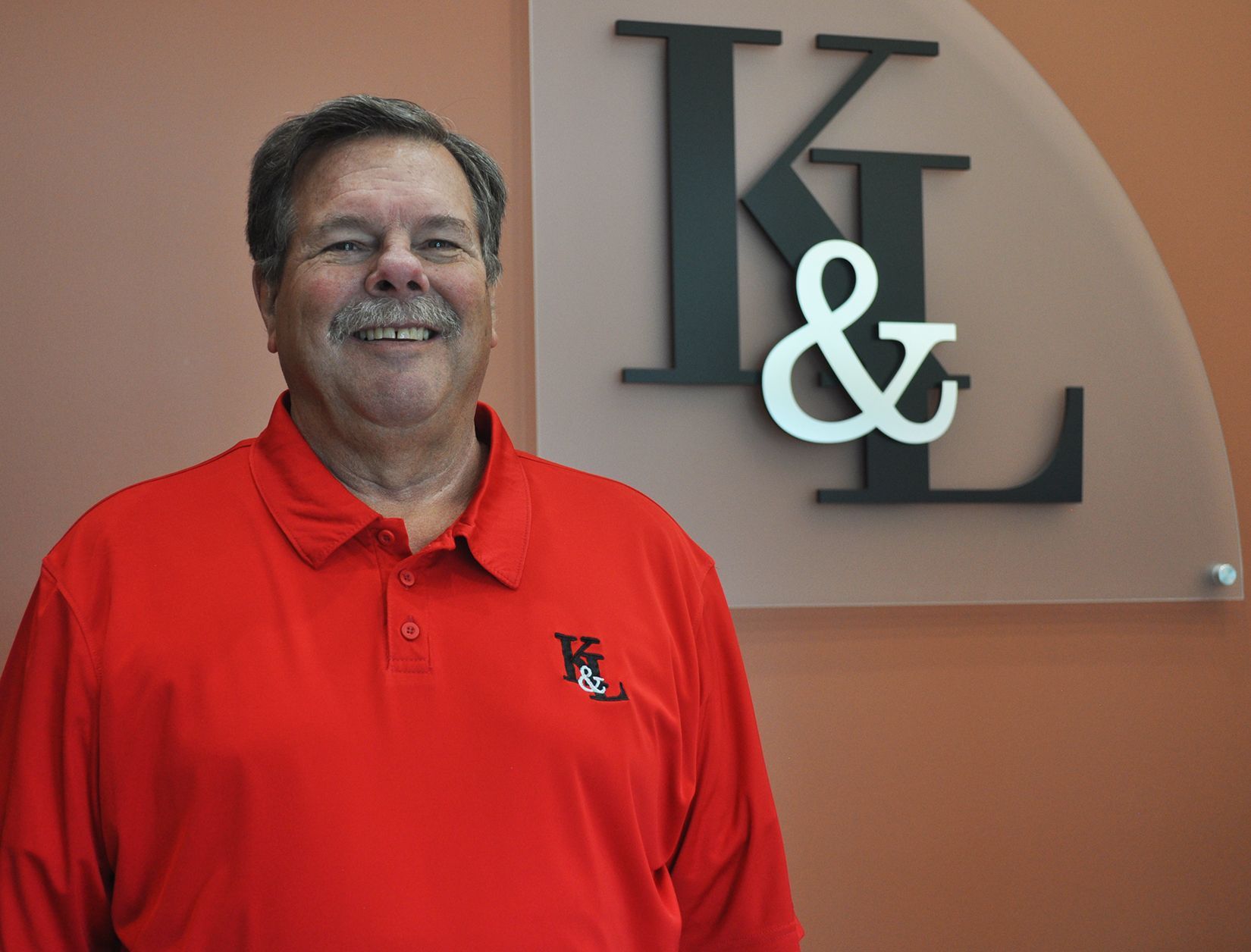 A man in a red shirt is smiling in front of a sign that says k & l