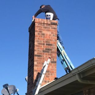 Worker on a ladder repairing a brick chimney on a roof under a clear blue sky