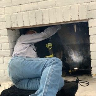 Person crouches in a fireplace opening, inspecting the dark interior with a flashlight.