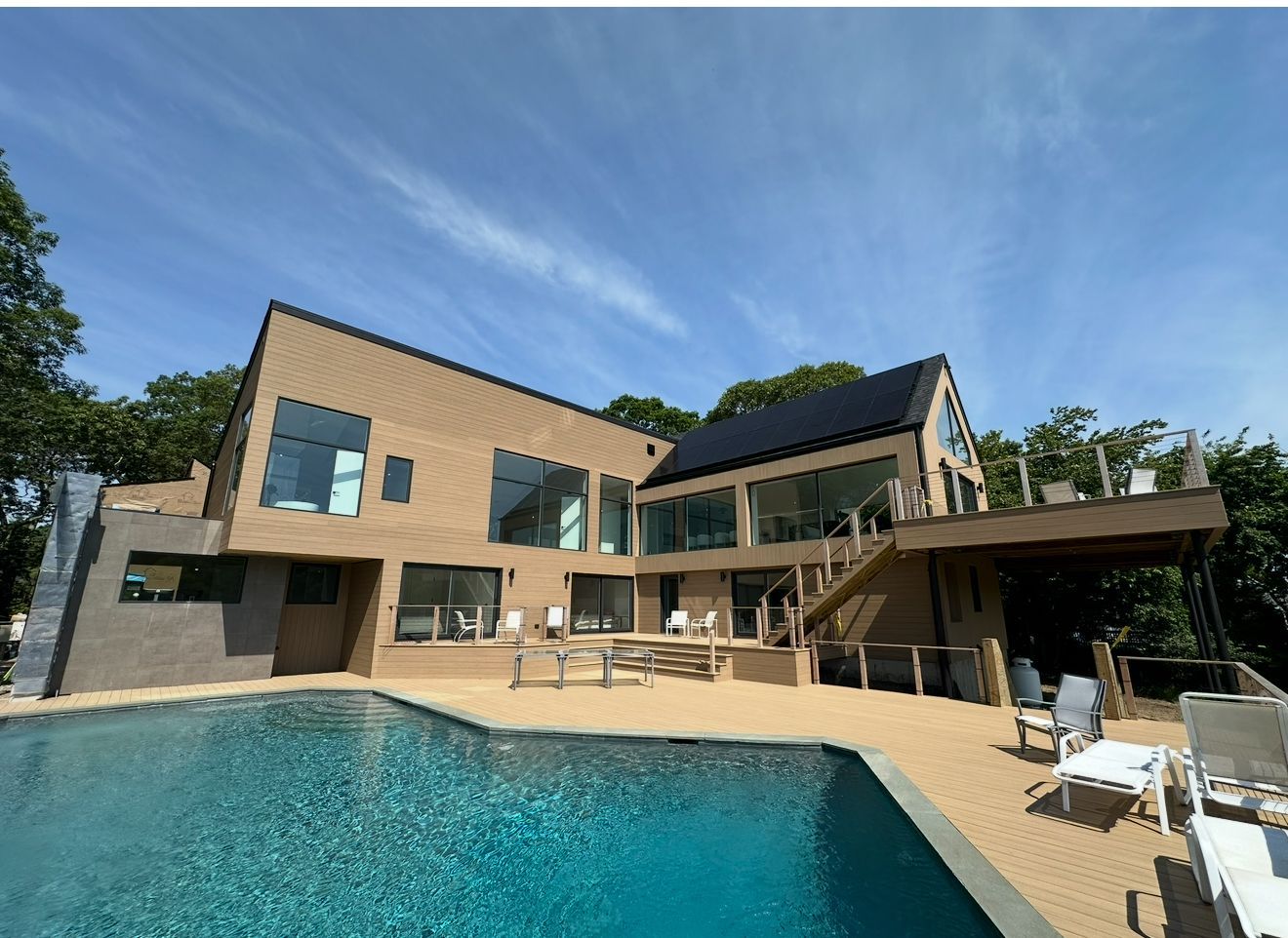 Modern house with a pool and solar panels on the roof. Light brown exterior with large windows, blue sky, and patio furniture.