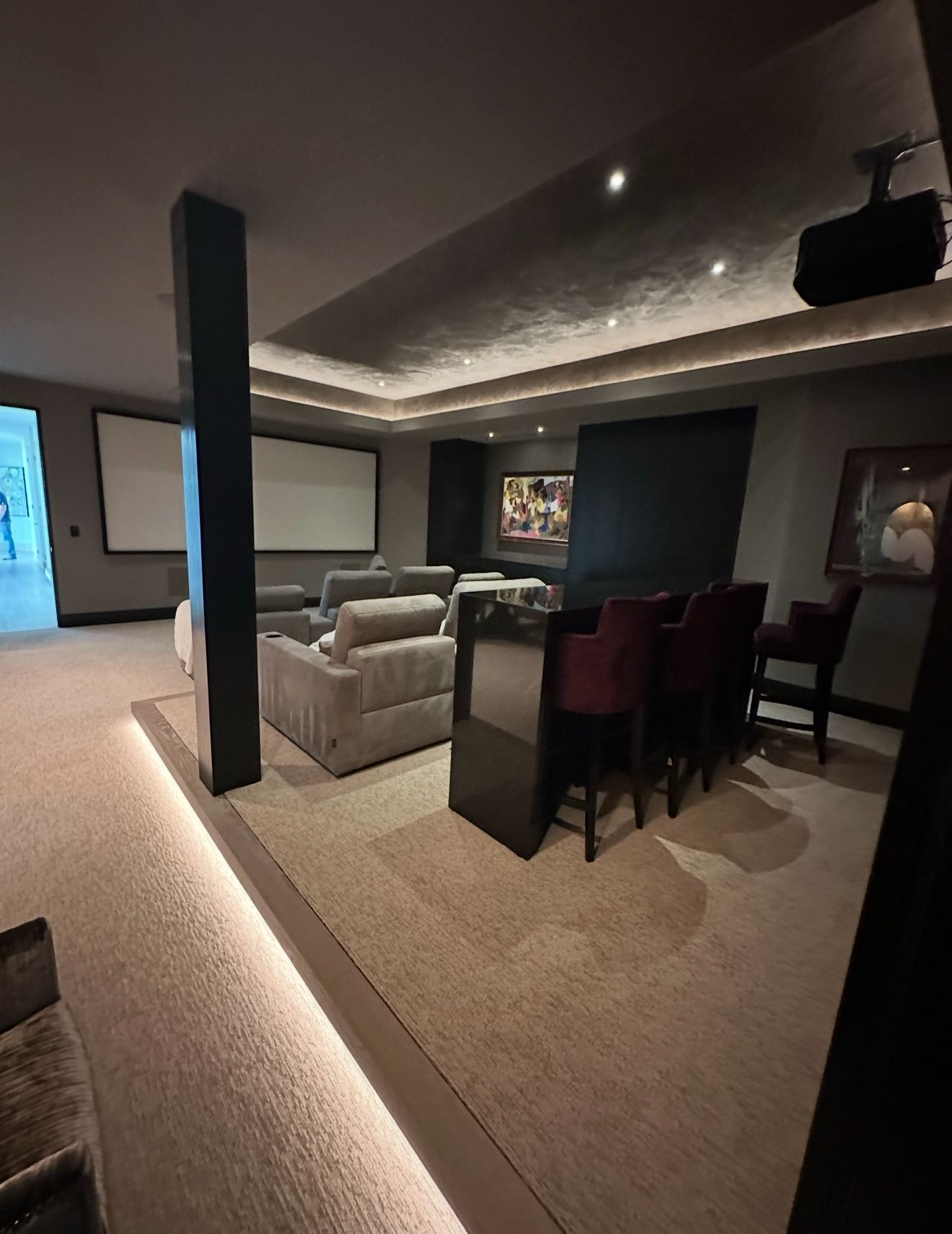 Luxurious home theater with a gray sectional sofa, bar seating, and two projection screens. Ambient lighting and dark accents create a sophisticated atmosphere.