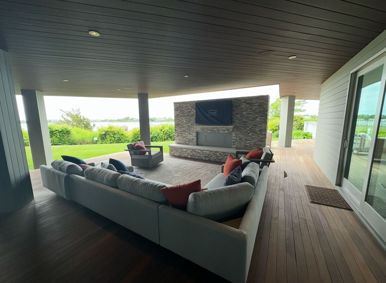 Outdoor living space with large sectional sofa, fireplace, and view of water. Deck with wood flooring and gray columns.